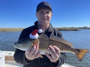 north-myrtle-beach-fishing-charters-redfish-red-drum-merry-christmas-gift man holding a redfish with a Santa hat on. Merry Christmas from North Myrtle Beach Fishing Charters